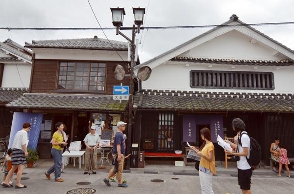 奥には日本家屋が2軒並んでいて、その手前で地図を持った男女が歩いている写真